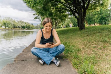 Mutlu gülümseyen 40 'lı yaşlardaki kadın beton şehir nehir kıyısında otururken akıllı telefon kullanıyor..