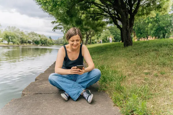 Mutlu gülümseyen 40 'lı yaşlardaki kadın beton şehir nehir kıyısında otururken akıllı telefon kullanıyor..