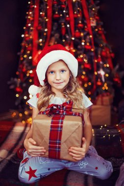 Pijamalı küçük bir kız, Noel Baba şapkalı bir kız şömine ve Noel ağacının yanında otururken hediye kutusu tutuyor. Mutlu Noeller. Mutlu yıllar. Noel süslemeleri olan rahat bir daire. Mucize zamanı.