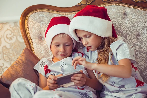 サンタの帽子とクリスマスのパジャマの小さな男の子と女の子は豪華なソファに座ってスマートフォンを見ています 新年のコンセプト — ストック写真