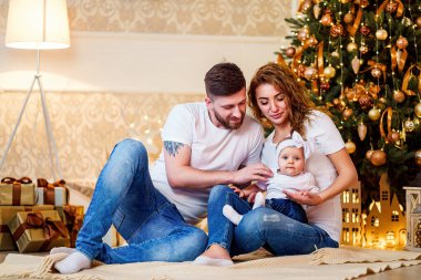 Mutlu bir aile. Genç ebeveynler, dekore edilmiş oturma odasında Noel ağacının yanında otururken küçük kıza sarılıyorlar. Noel 'i kutlamak. 