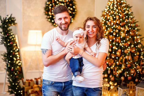 Happy young family standing near Christmas tree in living room while holding little cute baby girl in decorated interior. Celebrating Christmas.