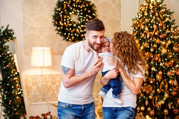 Happy family. mother and father hugging and kissing little baby girl in front of Christmas tree in decorated living room. Celebrating Christmas.