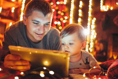 Babasıyla küçük oğlu Noel arifesinde sıcak ve rahat oturma odasında şöminenin yanında tablet ve Noel ağacı kullanıyor. Aile ve çocukla evde kış akşamı