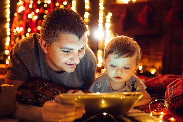 Father and little son using a tablet pc at home by a fireplace and Christmas tree in warm and cozy living room on Christmas eve. Winter evening at home with family and kid