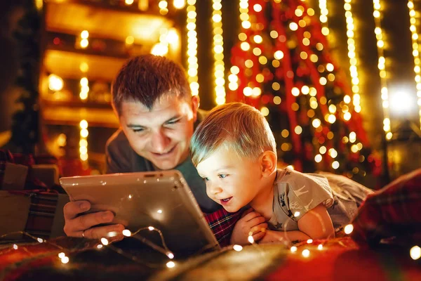 Father and little son using a tablet pc at home by a fireplace and Christmas tree in warm and cozy decorated living room on Christmas eve. Winter evening at home with family and kid