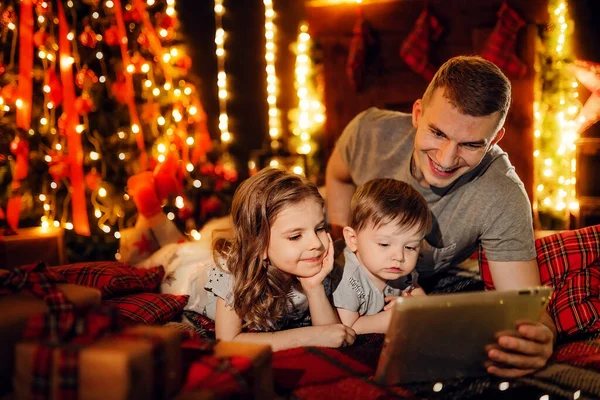 Father and his little daughter and son using a tablet pc at home by a fireplace in warm and cozy living room on Christmas eve. Winter evening at home with family and kids.
