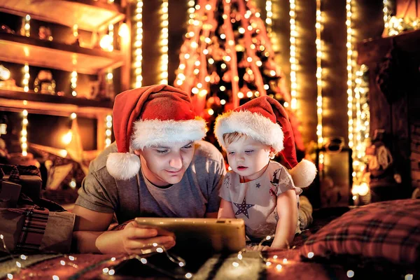 パジャマ姿の息子と家に寝そべっているサンタの帽子を持つ幸せな父親は 寝る前にデジタルタブレットコンピュータを使用しています 背景にクリスマスツリーとライト — ストック写真