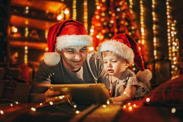 パジャマ姿の息子と家に寝そべっているサンタの帽子を持つ幸せな父親は 寝る前にデジタルタブレットコンピュータを使用しています 背景にクリスマスツリーとライト — ストック写真