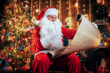 Noel Baba, Noel ağacının yanındaki koltukta oturuyor ve iyi çocukların listesini dikkatlice okuyor. Noel Zamanı. 