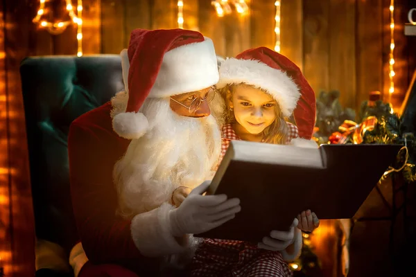 サンタ 句は暖炉の近くに座っている間サンタの帽子のかわいい女の子にクリスマスのおとぎ話を読みます 新年のコンセプト クリスマスタイム — ストック写真
