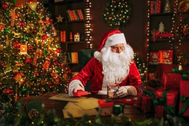 Noel Baba hediyeleri paketlemeye hazırlanırken evdeki Noel ağacının yanındaki masada oturuyor. Yeni yıl konsepti