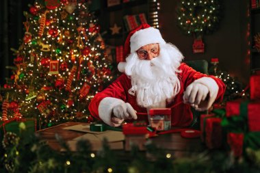 Noel Baba, Noel ağacının yanındaki masada otururken hediyeleri dikkatlice paketliyor. Yeni yıl konsepti