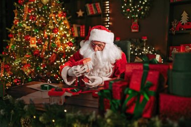 Noel Baba hediyelerini paketlerken evdeki Noel ağacının yanındaki masada oturuyor. Yeni yıl konsepti