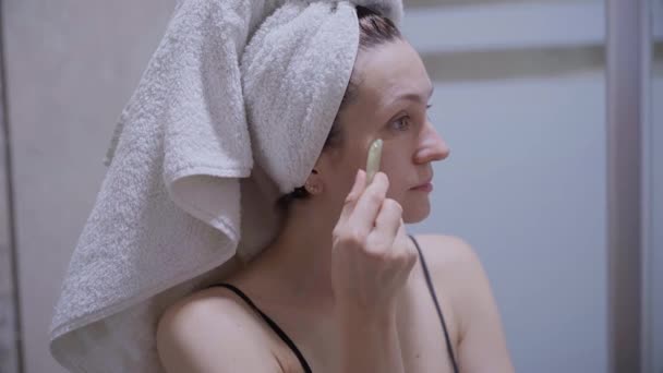 女人用刮胡刀做面部按摩 照镜子 她穿着睡衣 头上披着毛巾 光着肩膀 — 图库视频影像