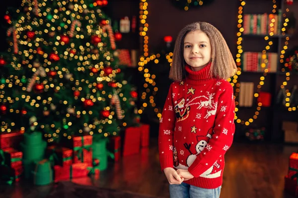 Little beautiful girl in Christmas sweater standing near Christmas tree. Portrait of cute girl in the New Year decorations.