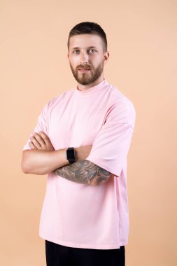 Modaya uygun sakallı yakışıklı hippi, şık saç kesimi ve modaya uygun giyinmiş, pembe tişörtlü, bej renkli stüdyo arka planında çapraz elleri olan yakışıklı hippi..