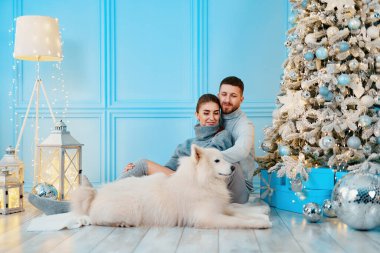 Mutlu çift evde oturup Noel ağacının yanında sarılıyor. Ocak, ev kıyafetleri ve kazaklar. konfor ve kalplerin sıcaklığı