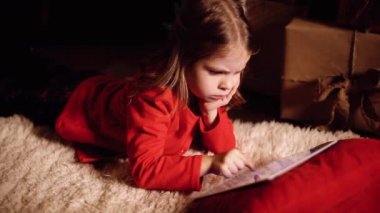 Evdeki Noel hediyelerinin yanında halıya uzanmış küçük bir kız tabletle oynamaya odaklanmış. Kamera hareket ediyor. 