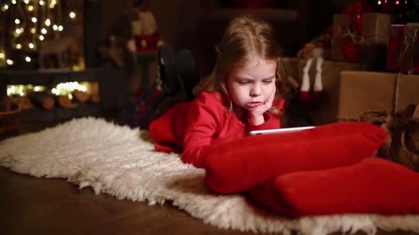 クリスマスプレゼントの近くのカーペットの上に横になり タブレットを使用して集中している少女 カメラの動き — ストック動画