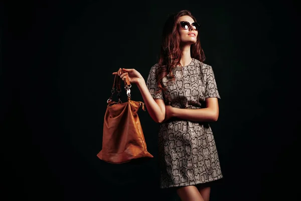 美丽和时尚 戴着太阳镜的时髦年轻女子 头戴蛇纹服装 手持黑色背景的棕色手提包 — 图库照片