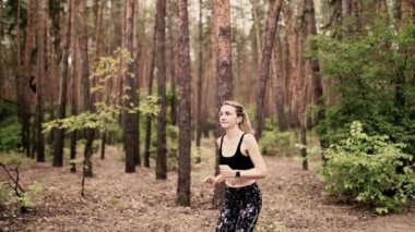 Güzel sarışın genç bir kadın spor kıyafetleri ve kulaklıklar takıp ormanda koşuyor. Yazın sağlıklı kalmak. 4 bin. Ağır çekim. Kamera hareket ediyor 