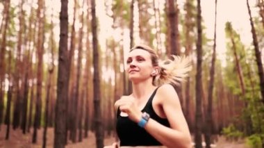 Güzel sarışın kız spor kıyafetleri ve kulaklıklarla ormanda koşuyor. Yazın sağlıklı kalmak. 4 bin. Ağır çekim. Kamera hareket ediyor.