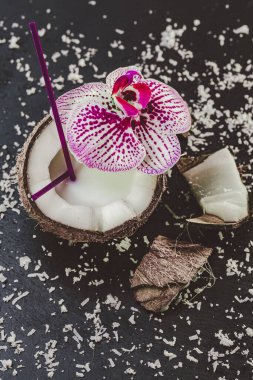 Taze hindistan cevizinin yukarıdan görünüşü, taştan siyah masa üzerine orkide ile süslenmiş samanla. Tropik meyve 