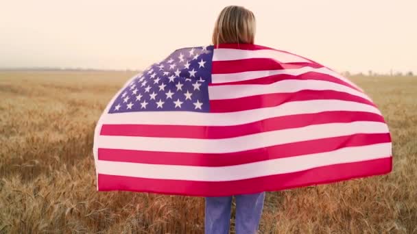 コムギ畑の真ん中にいるアメリカ国旗を胸に愛国的な幸せな女性の姿をバックにカメラに向ける 4Kだ スローモーション 愛国的概念 — ストック動画