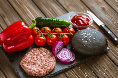 Siyah bir tahtanın üzerinde sığır burger, çiğ sığır eti köftesi, soğan, marul, domates, ketçap, dolma biber. Tahta masa. 