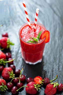 Yukardan bakıldığında taze çilek suyu ya da bardakta saman ve siyah masada taze çilek ve kirazlı smoothie görünüyor. Seçici odaklanma. Vejetaryen yemeği. Sağlıklı yiyecekler. Vitamin yiyeceği. 