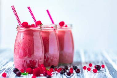 Front view of three berries milkshakes in glasses jars with straws with ingredients blueberries, raspberry, currant on wooden table. Selective focus. Healthy eating. Vegetarian diet. Copy Space 