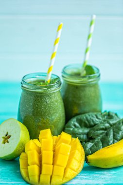 Freshly blended green smoothie with mango, apple, spinach and banana in glass jars with straws. Turquoise blue background. Selective focus. 