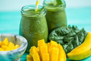 Closeup frreshly blended green smoothie with mango and banana in glass jars with straws. Turquoise blue background. Selective focus. 