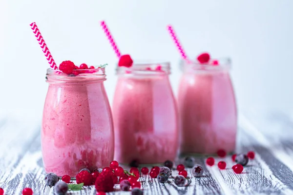 Front view of three berries milkshakes in glasses jars with straws with ingredients blueberries, raspberry, currant on wooden table. Selective focus. Healthy eating. Vegetarian diet.