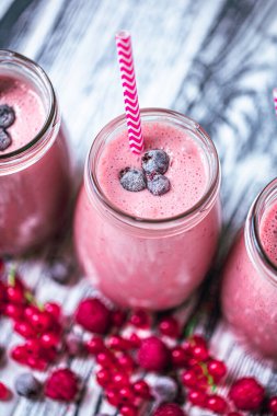 Top view of three berries milkshakes in glasses jars with straws which made from raspberry, blueberries, currant on wooden background. Selective focus. Healthy eating. Vegetarian food