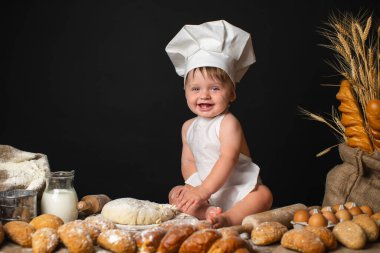 Happy little boy sits in a cook costume among a baking bread rolls of flour confectionery on the table on black background and kneading the dough. Space for text 