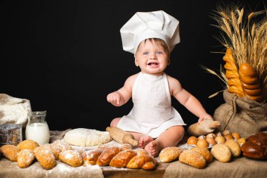Happy baby boy sits in a cook costume among a baking bread rolls of flour confectionery on the table on black background and holds rolling pin. Little chef kneading dough. Copy Space 