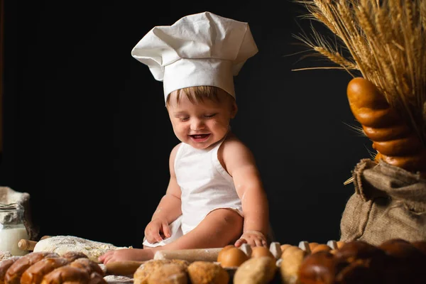 Laughing baby boy sits in a cook costume among a baking bread rolls of flour confectionery on the table on black background and holds rolling pin. Little chef kneading dough