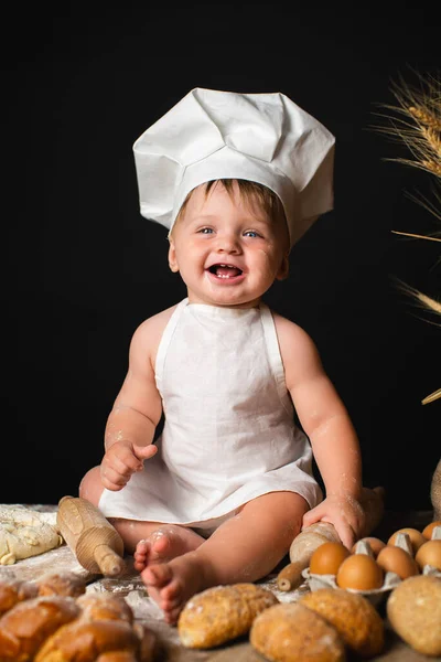 Cheerful little boy sits in a cook costume among a baking bread rolls of flour confectionery on the table on black background and holds rolling pin. Little chef kneading dough