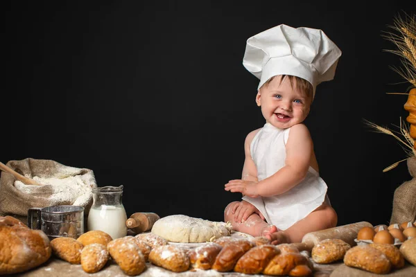 Funny little boy sits in a cook costume among a baking bread rolls of flour confectionery on the table on black background and kneading the dough. Little chef kneading dough. Space for text