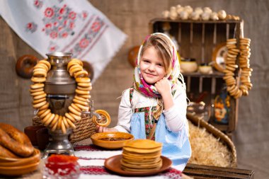 Cute little girl in a folk Russian headscarf and national dress with bunch of bagels and pancakes celebrating Maslenisa. Traditional Russian food for Shrovedite. clipart