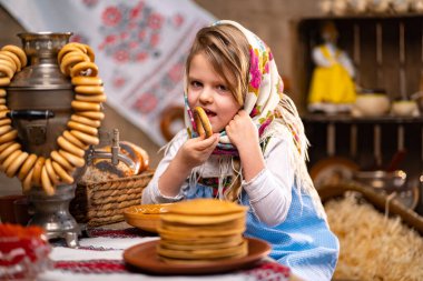 Cute little girl in a folk Russian headscarf and national dress eating bagels and celebrating Maslenisa. Traditional Russian food for Shrovedite. clipart