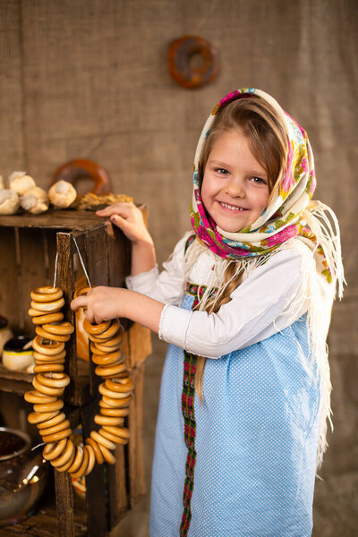 Улыбающаяся маленькая девочка в традиционном русском платке и национальном платье с бубликами. Крупный план портрета ребенка в народной одежде. Маслениса. Традиционная русская кухня.
