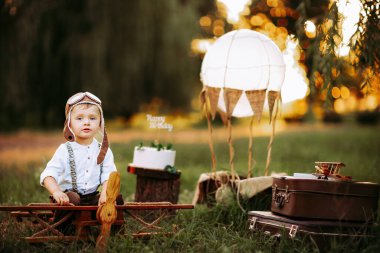Nice little pilot boy in vintage aviator hat looking at the camera outdoor while sitting on big toy wooden plane. Happy childhood. Dream come true 