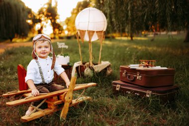 Dreaming little pilot boy in vintage aviator hat looking at the camera outdoor while sitting on big toy wooden plane. Happy childhood. Dream come true 