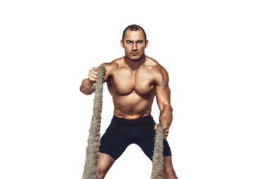 Strong young muscular man with naked torso doing exercises with rope while looking at camera white studio background. Copy Space 
