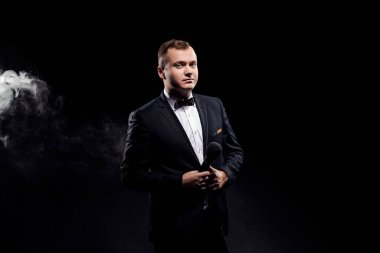 Elegant young man in black costume holding a microphone, posing against a dark background with smoke, actor, singer, show, host of the event. Space for advertising 