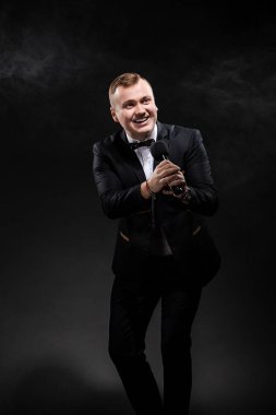 emotional man in black costume holding a microphone, posing against a dark background with smoke, actor, singer, show, host of the event.