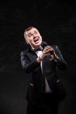 Portrait of young emotional man in black costume singing in microphone on black background, singer, actor, leading with microphone